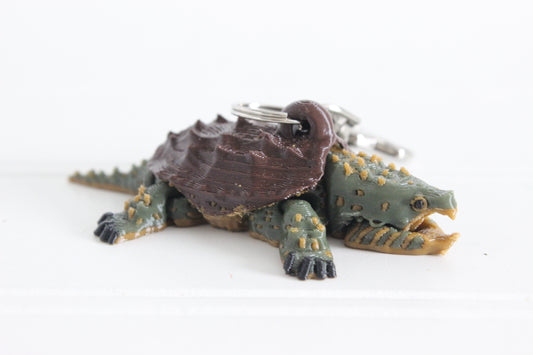 3D printed alligator snapping turtle keychain with dark brown shell and green textured body, lying flat on a white surface with swivel quick-clip key ring